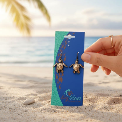 Big Blue by Roland St John bronze baby sea turtle drop earrings, detailed hatchling design, on display card on a beach.