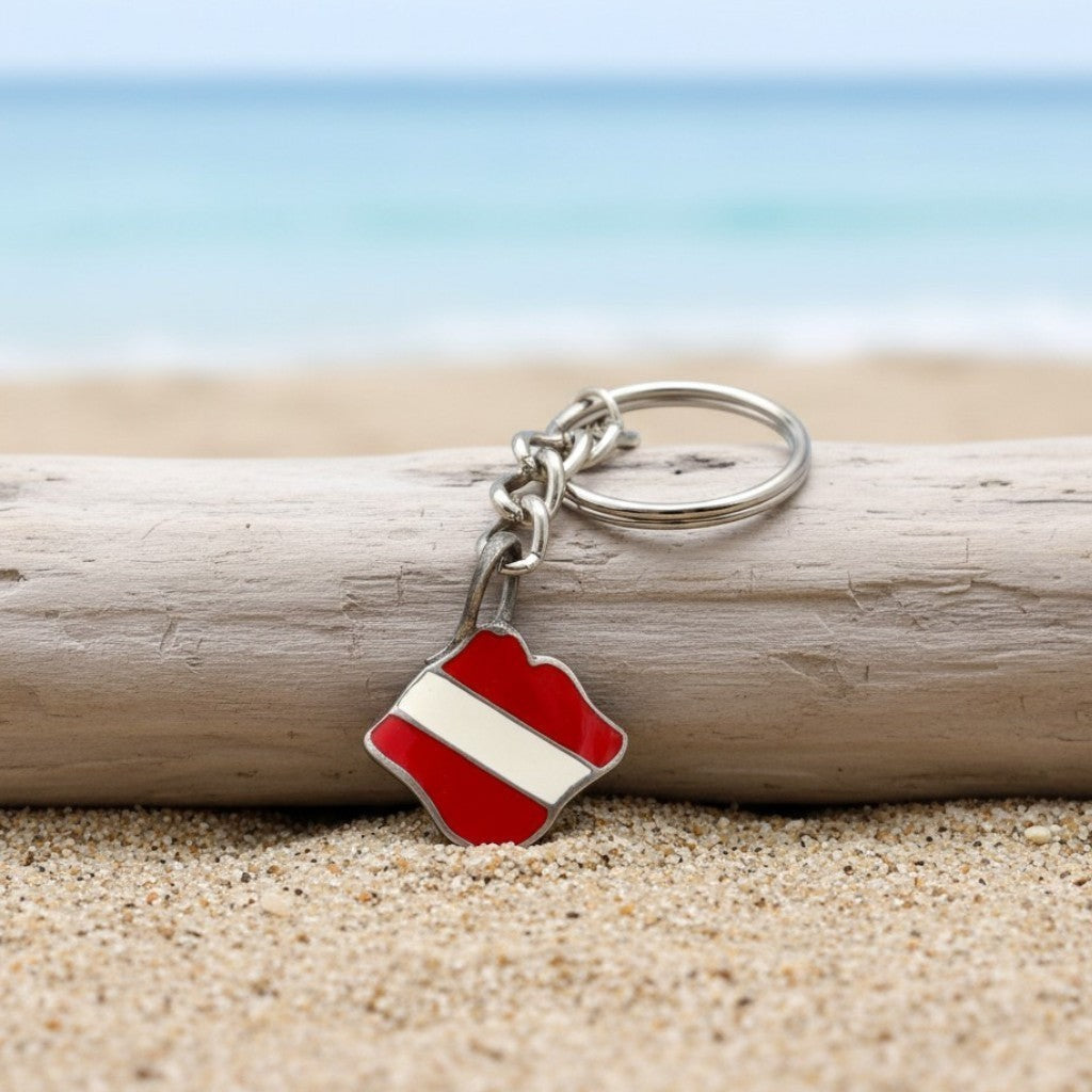 Big Blue by Roland St John pewter keychain with a red and white dive flag charm, set on a beach with driftwood and ocean background.