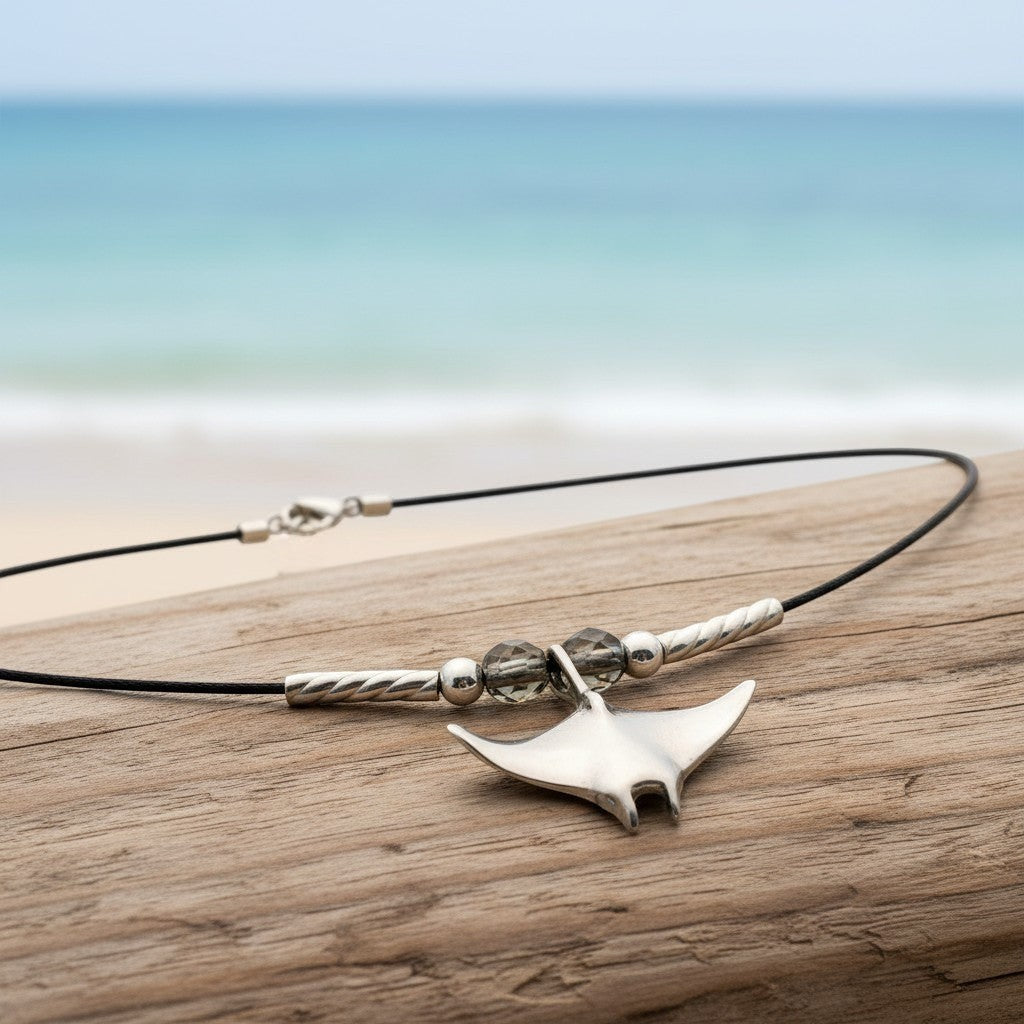 Big Blue by Roland St John sterling silver miniature manta ray choker necklace with grey beads on a black cord, set on driftwood with ocean background.