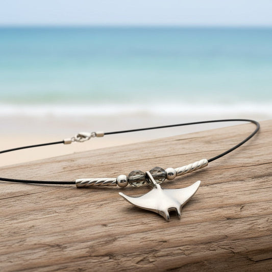 Big Blue by Roland St John sterling silver miniature manta ray choker necklace with grey beads on a black cord, set on driftwood with ocean background.