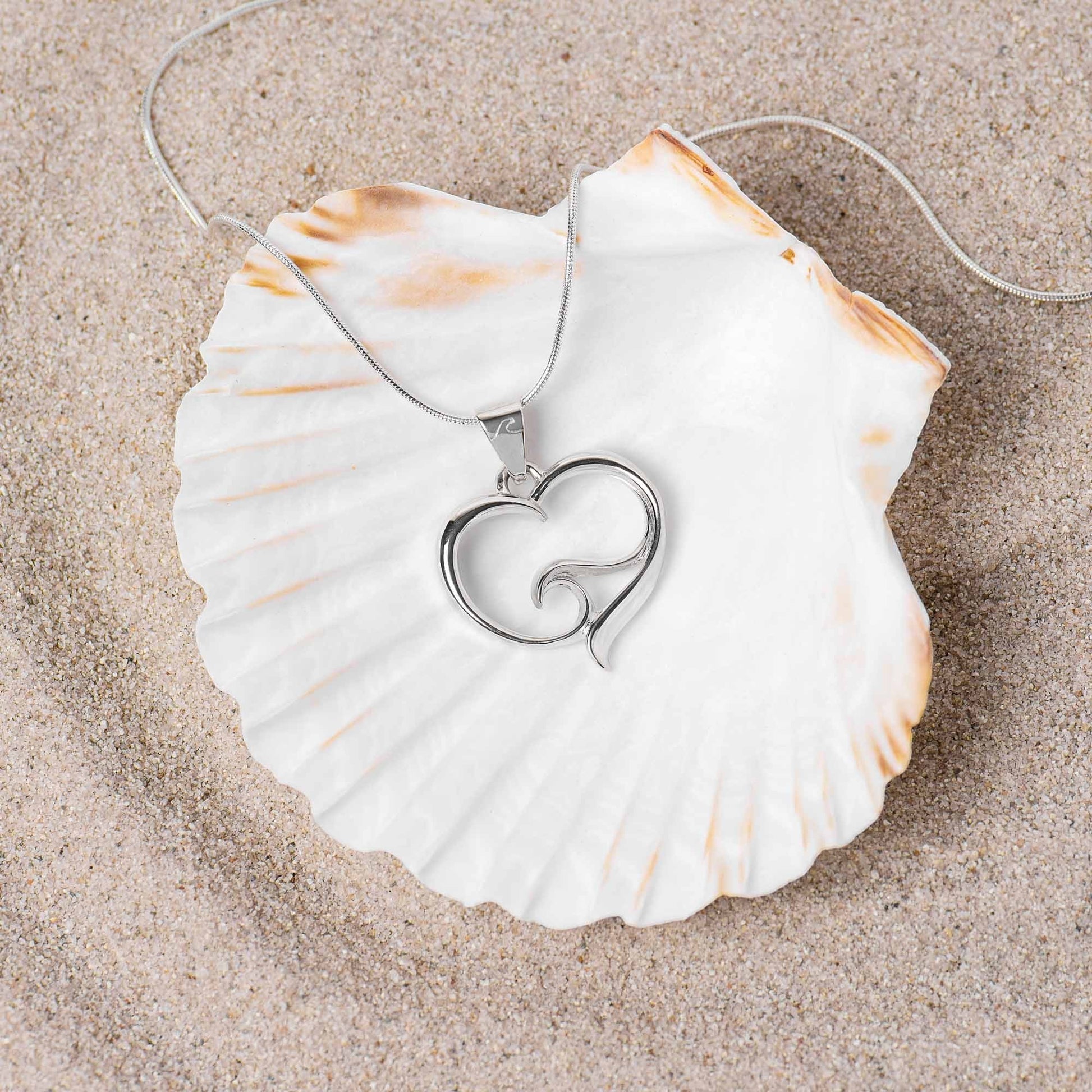 Big Blue by Roland St John sterling silver ocean wave heart pendant on snake chain, resting on a large seashell on sand.