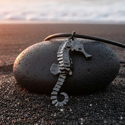 Big Blue by Roland St John jet black hematite seahorse pendant on black cord, resting on a dark stone on a black sand beach.