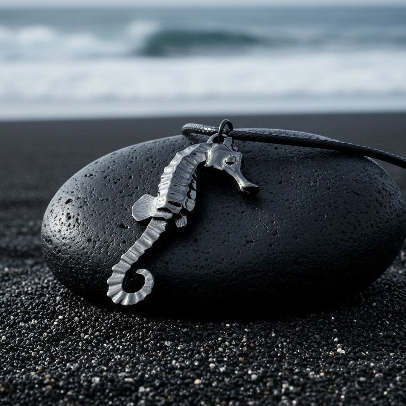 Big Blue by Roland St John jet black hematite seahorse pendant on black cord, resting on a dark stone on black sand beach.