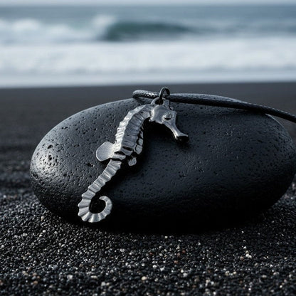 Big Blue by Roland St John jet black hematite seahorse pendant on black cord, resting on a dark stone on black sand beach.