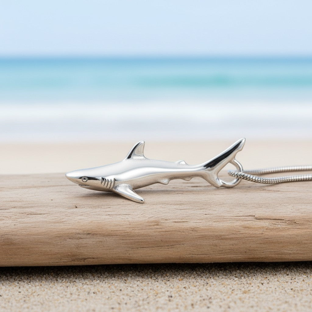 Big Blue by Roland St John realistic sterling silver grey reef shark pendant on a snake chain, resting on driftwood at the beach.