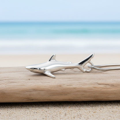 Big Blue by Roland St John realistic sterling silver grey reef shark pendant on a snake chain, resting on driftwood at the beach.