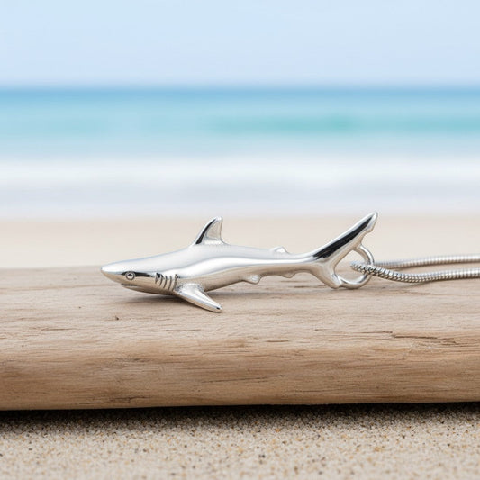 Big Blue by Roland St John realistic sterling silver grey reef shark pendant on a snake chain, resting on driftwood at the beach.