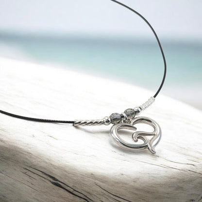 Big Blue by Roland St John sterling silver ocean wave heart choker necklace with grey beads on a black cord, set against a beach background.