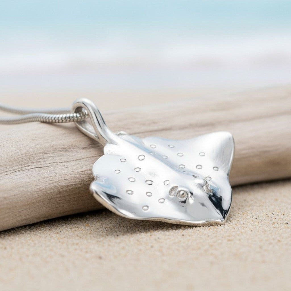 Big Blue by Roland St John sterling silver stingray pendant necklace with realistic details and a snake chain, set on a beach with driftwood.