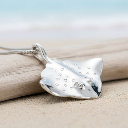 Big Blue by Roland St John sterling silver stingray pendant necklace with realistic details and a snake chain, set on a beach with driftwood.