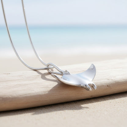 Big Blue by Roland St John sterling silver manta ray necklace with detailed wings, resting on driftwood by the sea.