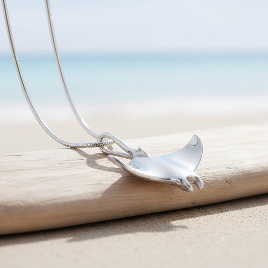 Big Blue by Roland St John sterling silver manta ray necklace with detailed wings, resting on driftwood by the sea.