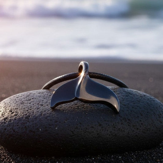Big Blue by Roland St John black whale tail pendant on a black cord, resting on a dark rock on a beach.