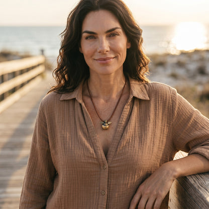 Big Blue by Roland St John bronze baby sea turtle necklace on a braided brown cord, worn by a woman on a boardwalk at sunset.