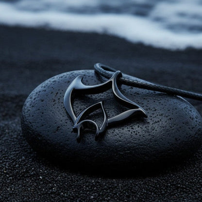 Big Blue by Roland St John Big Ray manta ray pendant necklace in jet black finish on a black cord, resting on a dark stone on black sand.