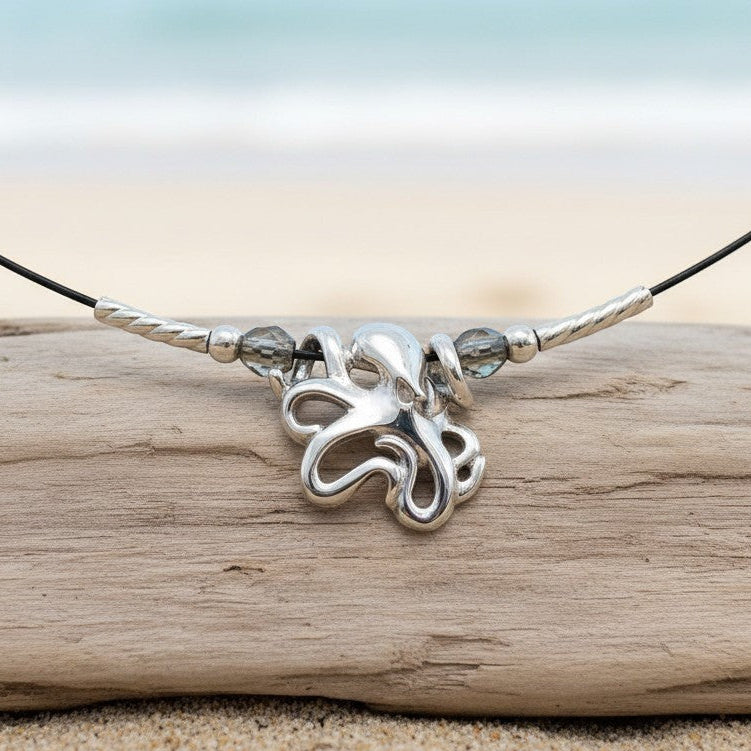 Big Blue by Roland St John sterling silver choker necklace with a mini octopus pendant and grey beads, displayed on a driftwood log on a beach.