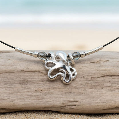 Big Blue by Roland St John sterling silver choker necklace with a mini octopus pendant and grey beads, displayed on a driftwood log on a beach.