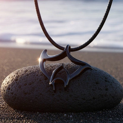 Big Blue by Roland St John Big Ray manta ray pendant necklace in jet black finish on a black cord, resting on a dark stone on a beach.