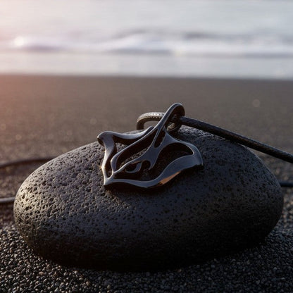 Big Blue by Roland St John jet black stingray pendant necklace on a dark stone on a beach.