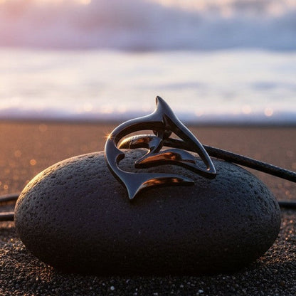 Big Blue by Roland St John "Frenzy" shark pendant necklace in jet black finish on black cord, displayed on a dark stone on a beach.