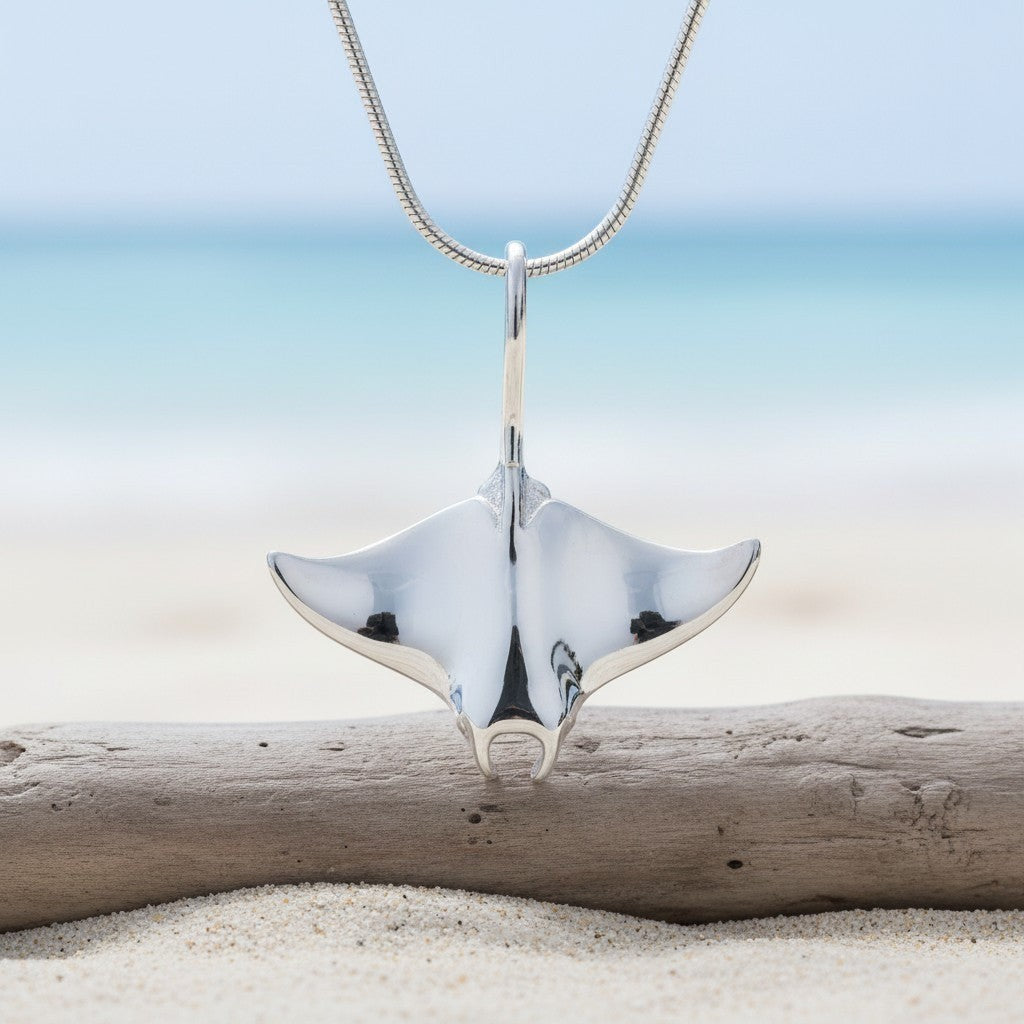 Big Blue by Roland St John realistic sterling silver manta ray pendant necklace on a snake chain, displayed on a beach with driftwood.