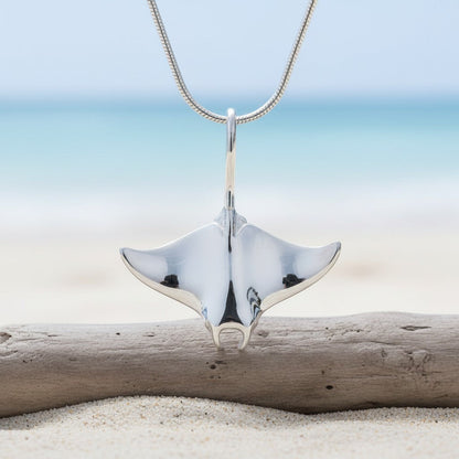 Big Blue by Roland St John realistic sterling silver manta ray pendant necklace on a snake chain, displayed on a beach with driftwood.