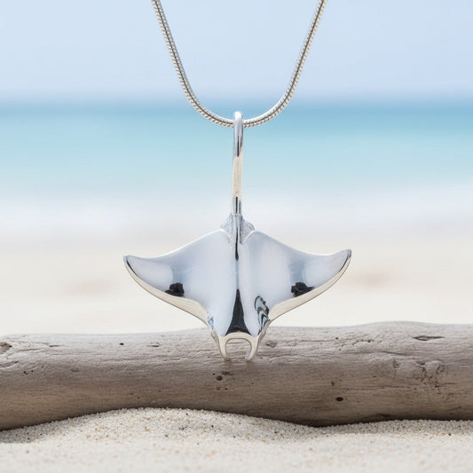 Big Blue by Roland St John realistic sterling silver manta ray pendant necklace on a snake chain, displayed on a beach with driftwood.