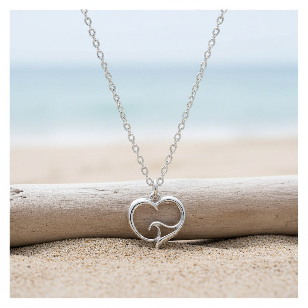 Big Blue by Roland St John sterling silver ocean wave heart necklace on a cable chain, resting on driftwood on a sandy beach.