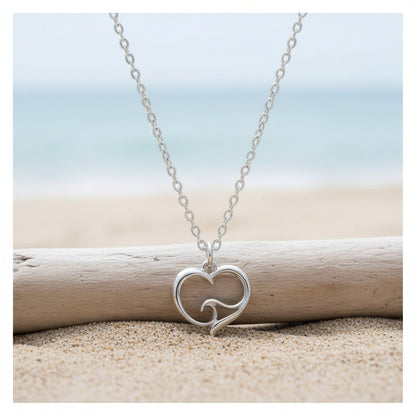Big Blue by Roland St John sterling silver ocean wave heart necklace on a cable chain, resting on driftwood on a sandy beach.
